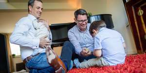 Pflegefamilie: Zwei Männer sitzen mit zwei Kindern auf einem Teppich