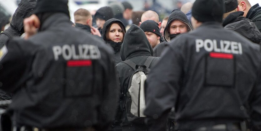 Polizisten stehen mit dem Rücken zur Kamera, dahinter eine Gruppe von Neonazis