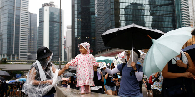 Demonstranten mit Regenschirmen führen ein kleines Mädchen über eine schmale Mauer