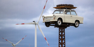 Im Hintergrund zwei Windräder, im Vordergrund ein Trabbi auf einem Masten weit über der Erde