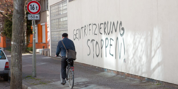 Ein Radfahrer vor einem schwarzen Graffiti an einer Wand in Kreuzberg, das sagt: "Gentrifizierung stoppen"
