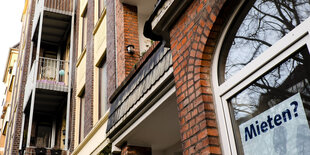 Ein Häuserfassade von unten fotografiert, in einem Fenster hängt ein Schild: "Mieten?"