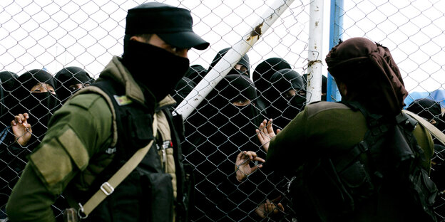 Zwei Männer in Uniform stehen vor einem Zaun, hinter dem einige Frauen in Burka stehen