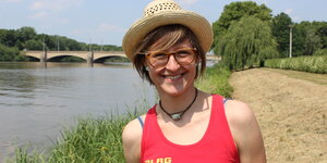 Eine junge Frau mit Hut und roten Oberteil steht vor einem Fluss