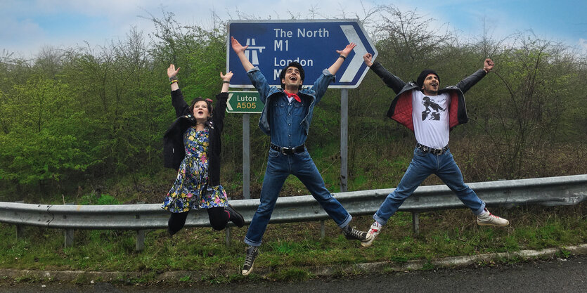 Drei Jungen springen mit ausgebreiteten Armen und Beinen in die Luft, vor einer Autobahnplanke