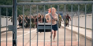 Zwei Frauen rennen über eine Brücke auf einen Zaun zu, sie werden von einer Zombiehorde verfolgt