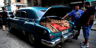 Ein Auto aus den 50er Jahren steht mit geöffneten Kofferraum in einer Straße. Im Kofferraum sind Zwiebeln zu erkennen, Männer stehen davor.