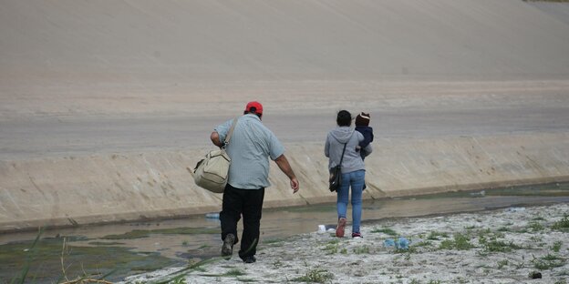 Eine Frau mit einem Kind auf dem Arm und ein Mann mit Reisetasche, sie laufen in den Bildmittelpunkt, wir sehen sie von hinten in einer kargen Landschaft