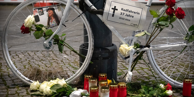 Ein weiß lackiertes Rad, mit Blumen verziert, davor Grablichter