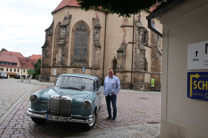 Ein hellblauer Mercedes, Baujahr 62. Daneben ein Mann