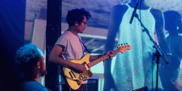 Janto Djassi Roessner spielt E-Gitarre. Im Hintergrund auf einer Leinwand ist ein junges Mädchen projiziert.