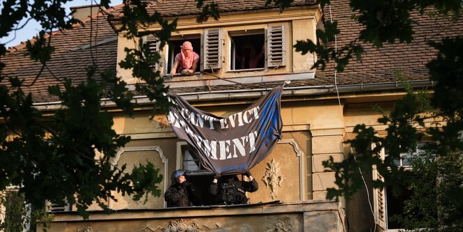 Ein vermummter Mensch hängt ein Transparent aus einem Balkon, einen Balkon weiter unten will es ein Polizist runter reißen