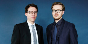 Zwei Männer in Anzügen stehen mit ernstem Gesichtsausdruck vor einem dunkelblauen Hintergrund