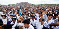 Hunderte Männer in weißen Hemden hocken auf dem Boden und strecken die Arme in die Luft