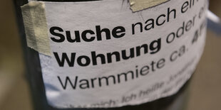 Auf einem Zettel steht "Suche nach... Wohnung...". Er ist mit Paketband an einer Laterne befestigt