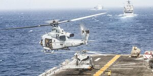 Ein Hubschrauber hebt gerade vom Deck eines Militärschiffs ab, ein anderer parkt, ein Lotse winkt