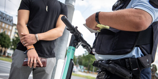 Auf dem Bild sind die Arme eines Mannes und die eines Polizisten in Uniform zu sehen. Sie stehen sich gegenüber, dazwischen steht ein E-Tretroller