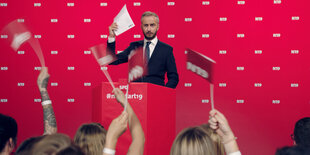 Ein Mann steht an einem kleinen Rednerpult und hält ein Formular in die Höhe. Es ist Jan Böhmermann. Vor ihm steht eine kleine Gruppe an Menschen und jubelt ihm zu