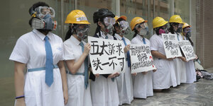 Schüler*innen knien vor einem Gebäude. Sie tragen Helme, Gasmasken und Plakate