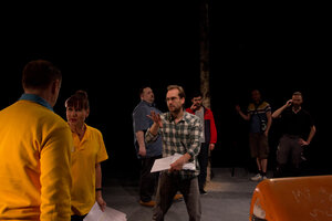 Ein Mann gestikuliert, umgeben von SchauspielerInnen in einem dunklen Theatersaal Ein Mann gestikuliert, umgeben von SchauspielerInnen in einem dunklen Theatersaal