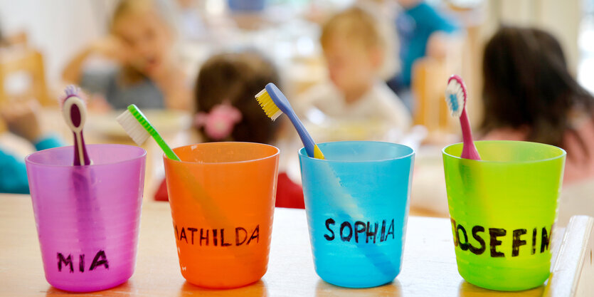 Vier verschiedenfarbige Zahnputzbecher stehen auf einem Tisch, in jedem steckte eine Zahnbürste, die Becher sind mit Mädchennamen beschriftet