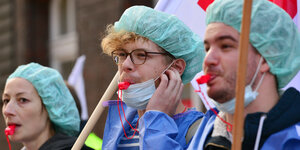 Drei Pfleger*innen protestieren in Kitteln und mit Trillerpfeifen.