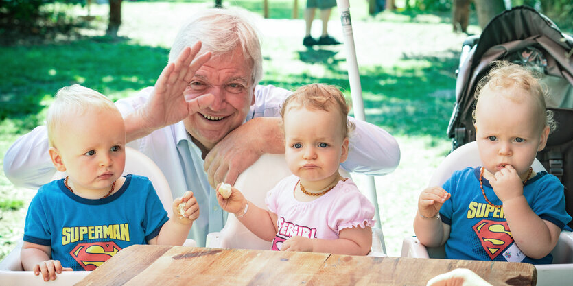 Volker Bouffier, Ministerpräsident des Landes Hessen, hockt winkend hinter den 14 Monate alten Drillingen Thies, Leonie und Bennett, die beim Drillingstreffen in ihren Hochstühlen sitzen.