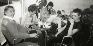 Sitzende Menschen um einen kleinen Cafétisch.Der noch junge Sänger Blixa Bargeld ganz in Schwarz. Im Hintergrund steht ein Gummibaum. Schwarzweißaufnahme aus dem Café Mitropa