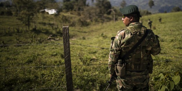 Ein Soldat in tarnfarbener Uniform steht vor grünen Hügeln, im Hintergrund wartet ein Transporthubschrauber
