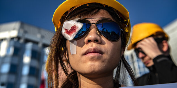 Eine Demonstrantin mit gelbem Helm und Sonnenbrille, auf dem rechten Auge eine Kompresse mit rotem Fleck