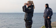 Eine blonde Polizistin in Uniform blickt mit einem Fernglas von der Insel Lesbos in Griechenland auf das Mittelmeer. Ein Polizist steht rechts im Hintergrund. Sein Blick ist ebenfalls aufs Meer gerichtet.