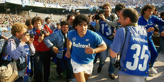 Diego Maradona läuft im blauen Trikot ins Stadium ein, er ist umringt von Fans und Journalisten