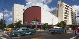 Ein großes Kinogebäude ragt zwischen zwei Hochhäusern in den blauen Himmel, davor fahren auf der Straße zwei Straßenkreuzer