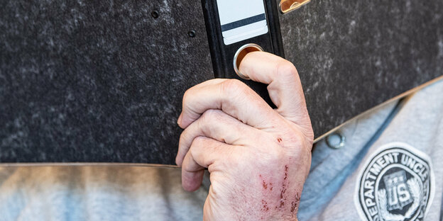 Der Angeklagte Andreas V. hält sich mit einer Hand, die wohl aufgrund einer Krankheit Kratzspuren aufweist, einen Aktenordner vor das Gesicht