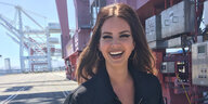 Lana Del Rey steht an einem Hafen. Im Hintergrund sind Lastenkräne. Del Rey lacht mit offenem Mund. Sie hat lange braune Haare und trägt eine Trainings-Jacke, die leicht geöffnet ist.
