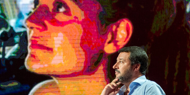 Matteo Salvini sitzt vor einer Leinwand, auf der das Gesicht von Carola Rackete zu sehen ist.