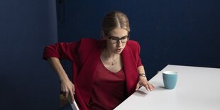 Eine Frau im roten Kostüm stützt sich beim Aufstehen auf die Stuhllehne und den Tisch