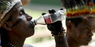 Ein Amazonas-Bewohner trinkt aus einer Coca-Cola-Plastikflasche. Er trägt bunten Federschmuck auf dem Kopf