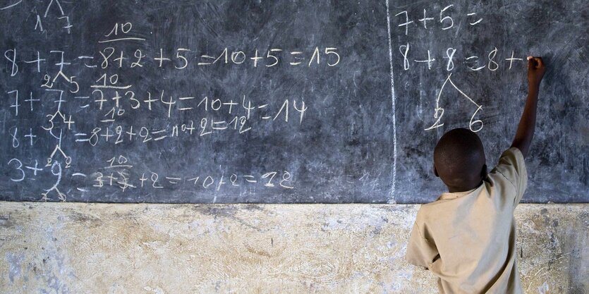 Man sieht einen etwa 6 Jahre alten Jungen von hinten, der eine Mathe-Aufgabe auf eine Schieferwand malt.