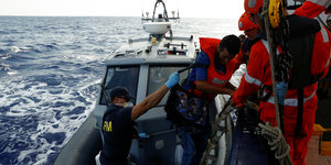 Ein maltesischer Soldat steht auf einem Militärschiff. Er trägt Mundschutz und Handschuhe und hilft einem Geflüchteten von Bord der Alan Kurdi über eine Treppe auf das Militärschiff zu gelangen. Der Geflüchtete trägt eine orangene Rettungsweste