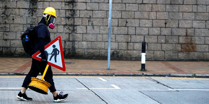 Eine Demonstrantin in Hongkong trägt ein Schild, auf dem vor einer Baustelle gewarnt wird. Die Person ist komplett schwarz gekleidet, trägt eine Athemmaske und einen gelben Helm.