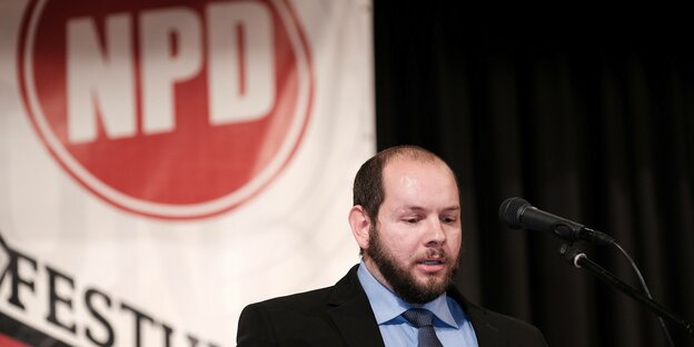Stefan Jagsch vor einem NPD-Schriftzug. Er spricht am Mikrofon
