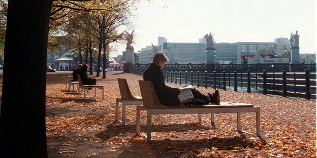 Eine Person sonnt sich im herbstlichen Berlin am Ufer der Spree auf einer Bank.