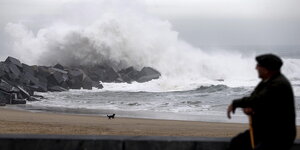 Ein alter Mann mit einer Baskenmütze blickt auf das stürmische Meer an einer Küste, wo sich hohe Wellen brechen.