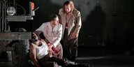 Zwei Männer mit blutverschmierter Kleidung beugen sich über einen verletzten Mann in Frank Castorfs Inszenierung von Verdis Oper "La forza del destino"