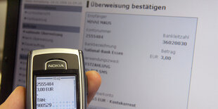 Handyscreen mit TAN vor Computerbildschirm, auf dem Überweisung zu sehen ist