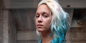 Eine junge Frau mit bläulich gefärbten Haaren schaut mit ernstem Blick in die Kamera