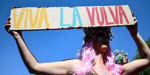 Person mit rosa Perücke hält ein Schild hoch. Darauf steht in bunter Schrift: "Viva la Vulva".