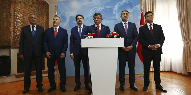 Sechs Männer stehen in dunklen Anzügen in einem Saal, einer von ihnen vorgerückt an einem Stehpult mit Mikrofon. Es ist der türkische Ex-Regierungschef Ahmet Davutoğlu.