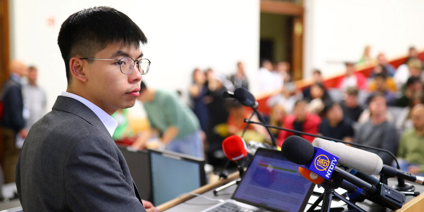 Joshua Wong steht in einem voll besetzten Hörsaal am Pult vor mehreren Mikros